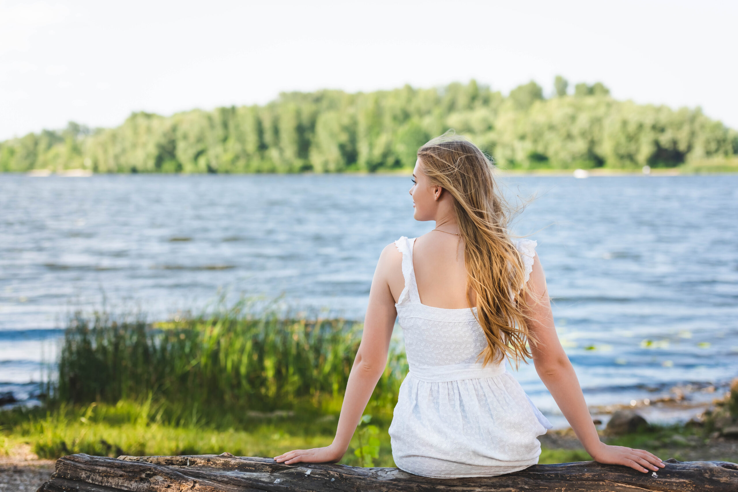back view of girl sitting on trunk of tree near river shore and looking away