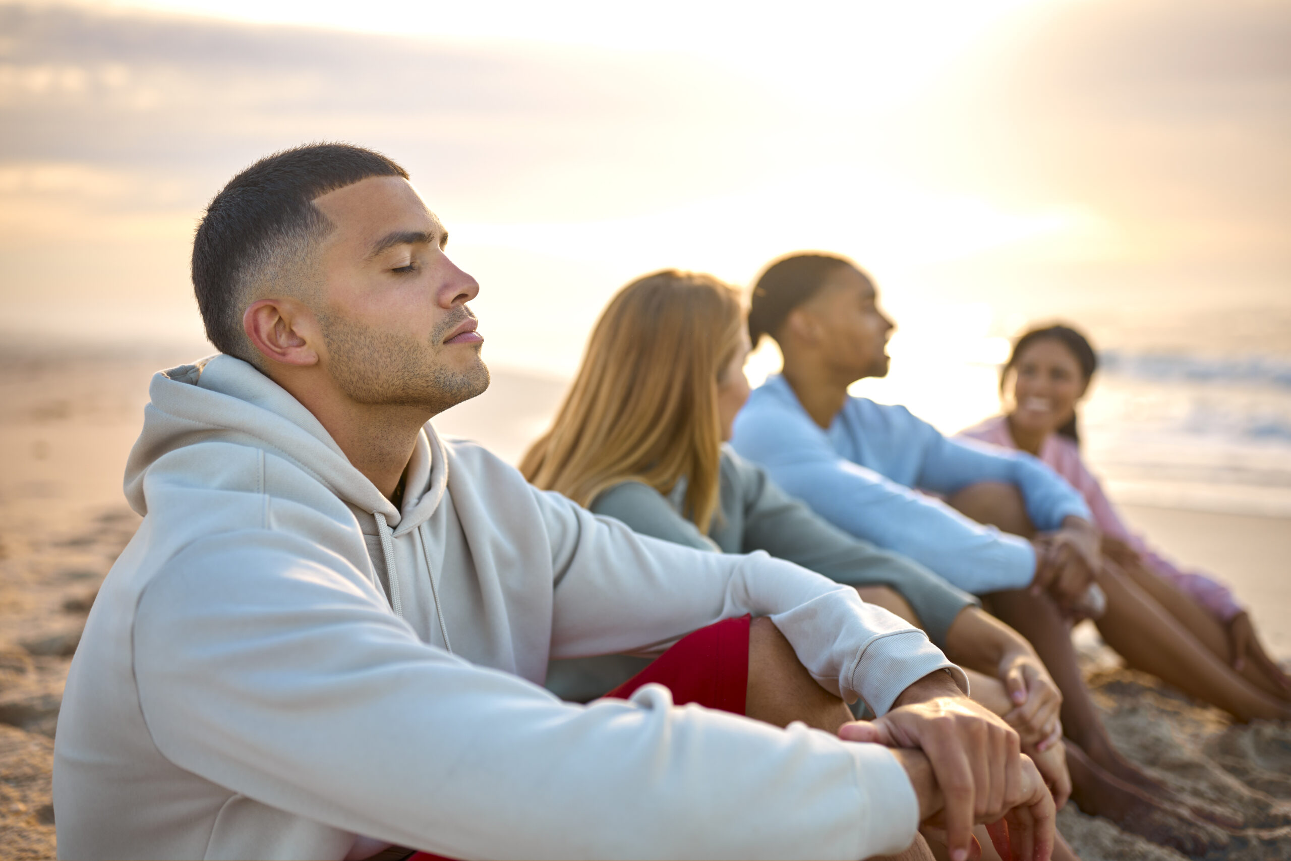 Man With Closed Eyes Relaxing With Friends On Vacation Sitting On Beach Watching Sunrise Together