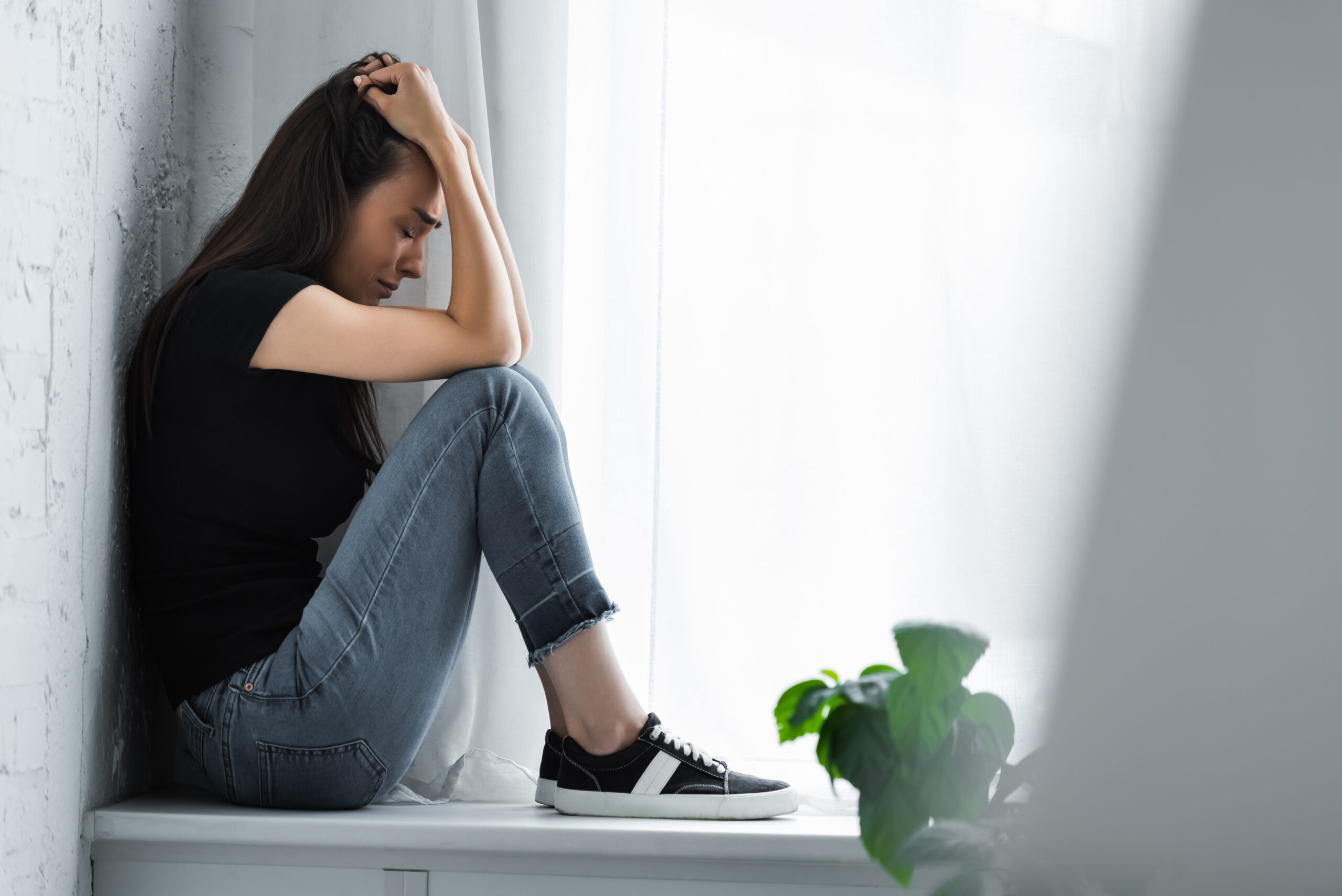 selective focus of young woman crying while sitting on window sill and suffering from depression ocd and addiction treatment